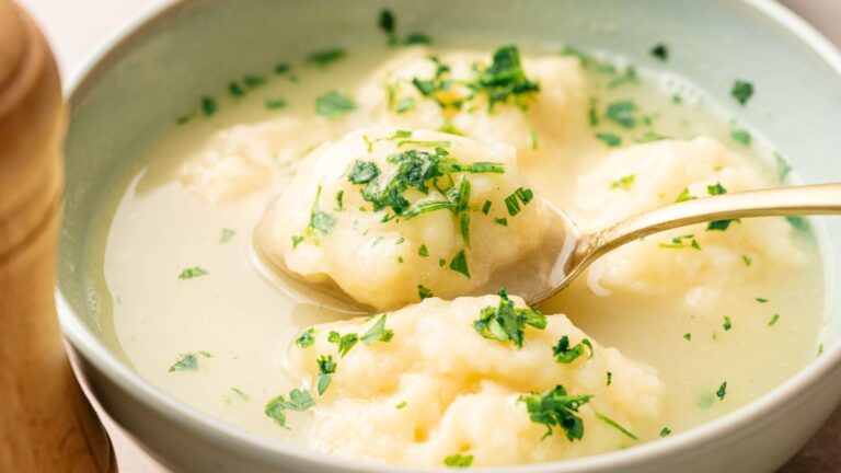 A bowl of soup with fluffy dumplings, garnished with chopped parsley, with a spoon lifting one dumpling from the bowl.