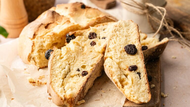 A loaf of rustic bread with raisins, partially sliced, sits on parchment paper with a few crumbs scattered around.