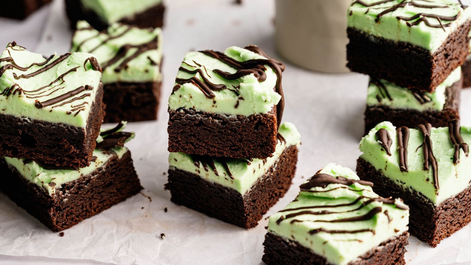 Brownie squares topped with a layer of green mint frosting and drizzled with chocolate, arranged on parchment paper.