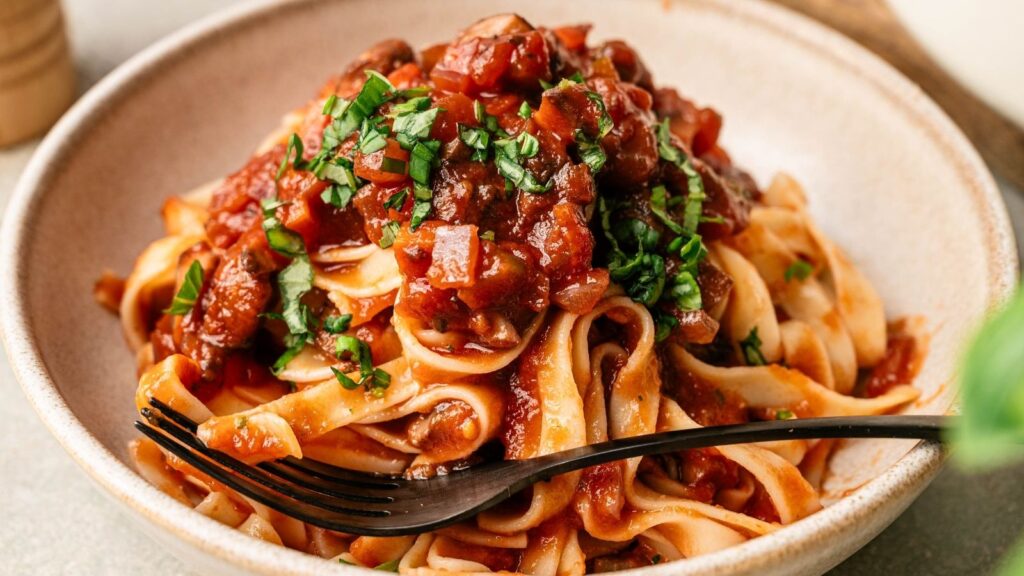 A bowl of tagliatelle pasta topped with tomato sauce, chopped herbs, and a black fork resting on the side.