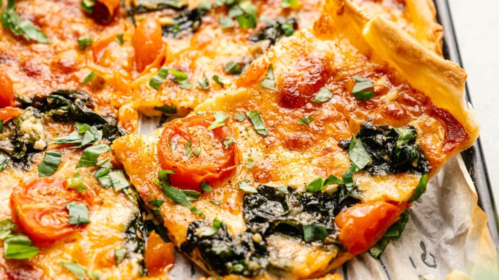 Close-up of a rectangular pizza topped with melted cheese, cherry tomatoes, spinach, and chopped fresh herbs on a baking tray.