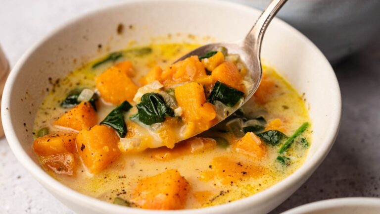 A bowl of creamy soup with chunks of orange squash, wilted spinach, and visible black pepper, with a spoon lifting a portion.