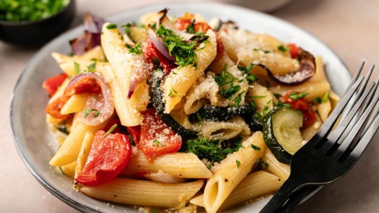 Plate of penne pasta with roasted vegetables, including zucchini, red onion, and tomatoes, garnished with grated cheese and chopped parsley, with a black fork on the side.