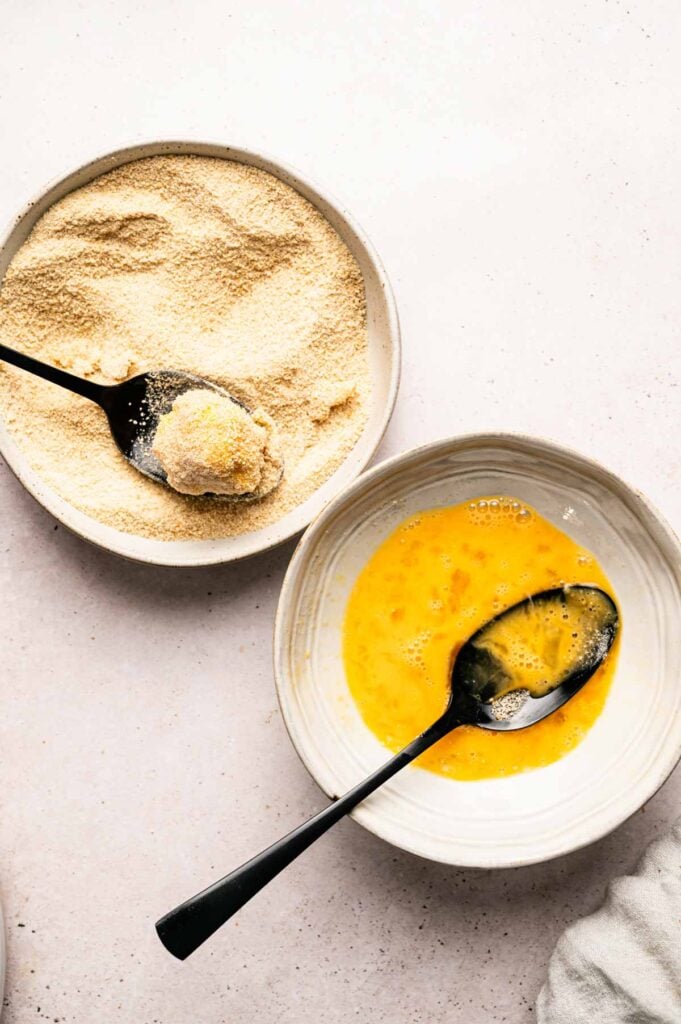Two bowls on a light surface: one with beaten eggs and a spoon, the other with breadcrumbs and a spoon holding a portion of breadcrumbs, prepared for coating Mac and Cheese Balls.