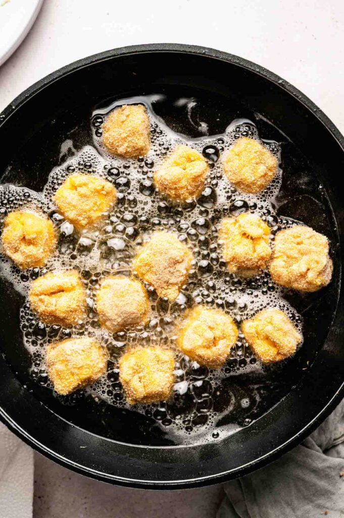Mac and Cheese Balls are being breaded and deep-fried in hot oil in a black frying pan.