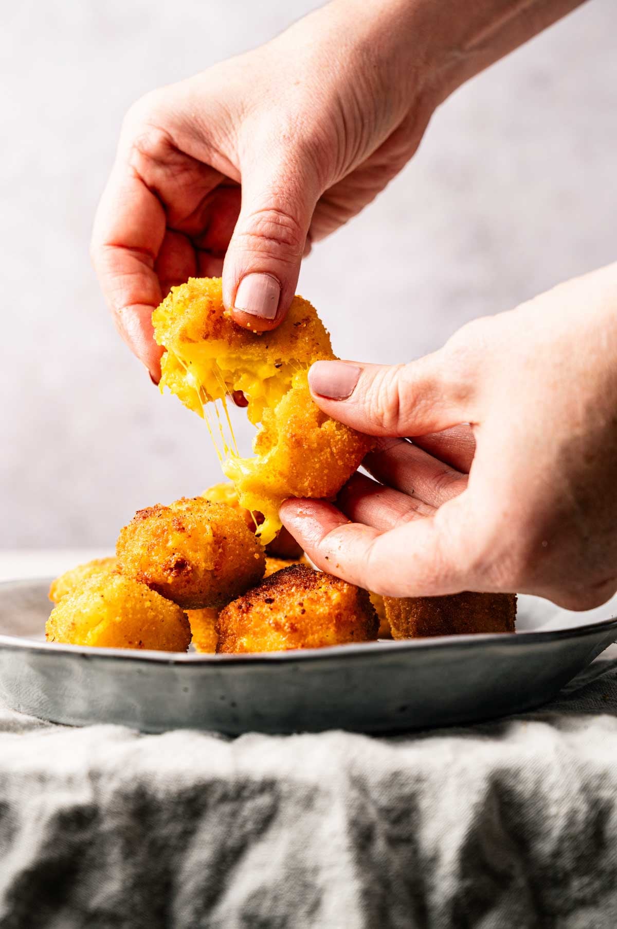 Hands pull apart a golden, breaded Mac and Cheese Ball with melted cheese stretching, above a plate filled with similar cheese balls.