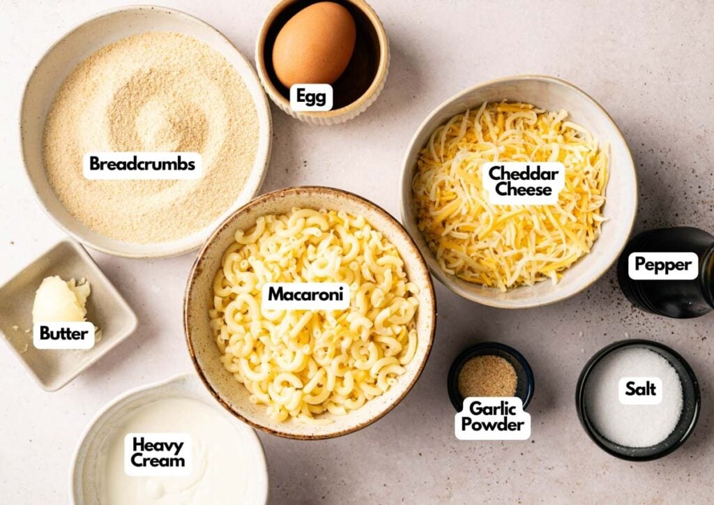 Bowls containing ingredients for Mac and Cheese Balls&mdash;breadcrumbs, egg, shredded cheddar cheese, macaroni, pepper, salt, garlic powder, heavy cream, and a pat of butter&mdash;are arranged on a light surface.