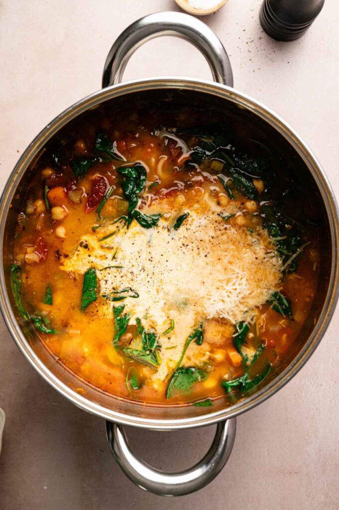 A pot of chickpea pasta soup is topped with a swirl of cream and grated Parmesan before the final stir.