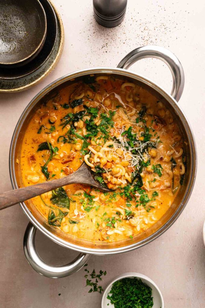 A pot of creamy Marry Me Chickpea Soup with tomato, spinach, beans, and grated cheese, garnished with chopped herbs, sits on a light surface next to stacked bowls and a small bowl of parsley.