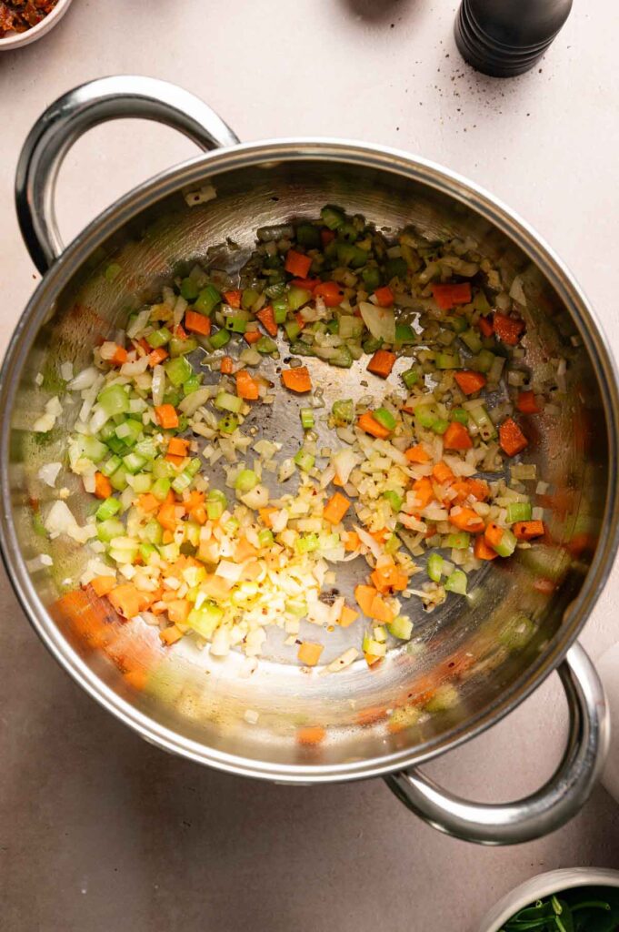Chopped onions, carrots, celery, and garlic are sautéing in a stainless steel pot on a light-colored countertop, creating the flavorful base for a comforting Marry Me Chickpea Soup.