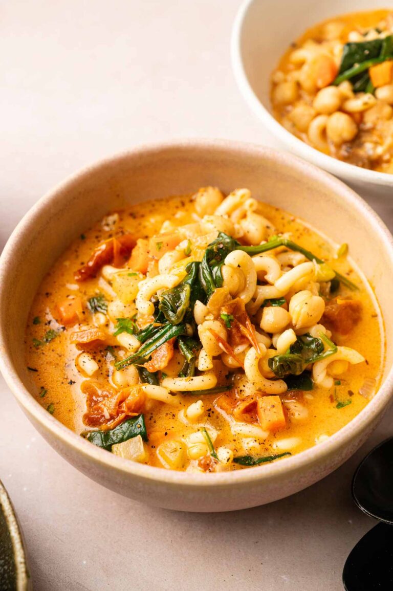 A bowl of Marry Me Chickpea Soup with creamy vegetable broth, pasta, greens, diced tomatoes, and chickpeas sits on a light surface.