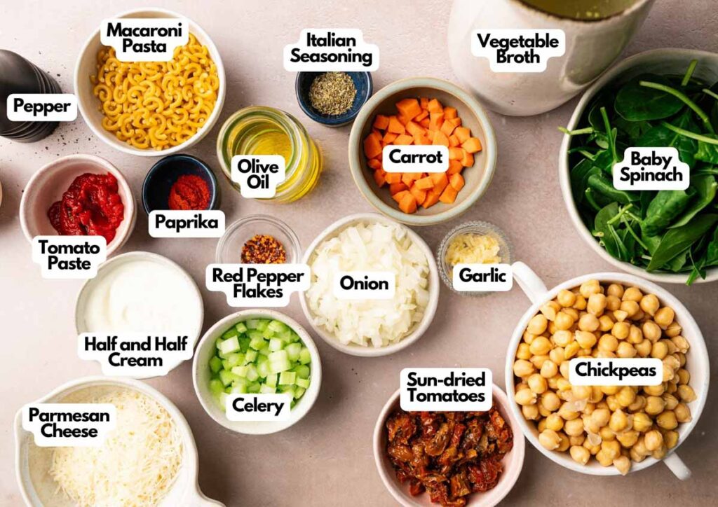 Top-down view of labeled ingredients for Marry Me Chickpea Soup in bowls on a countertop, including vegetables, pasta, chickpeas, seasonings, olive oil, cream, broth, and cheese.