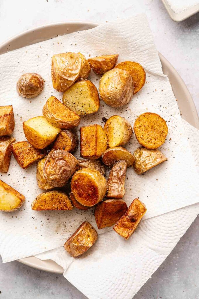 Golden, crispy potato pieces are arranged on a white paper towel atop a beige plate, lightly seasoned with salt