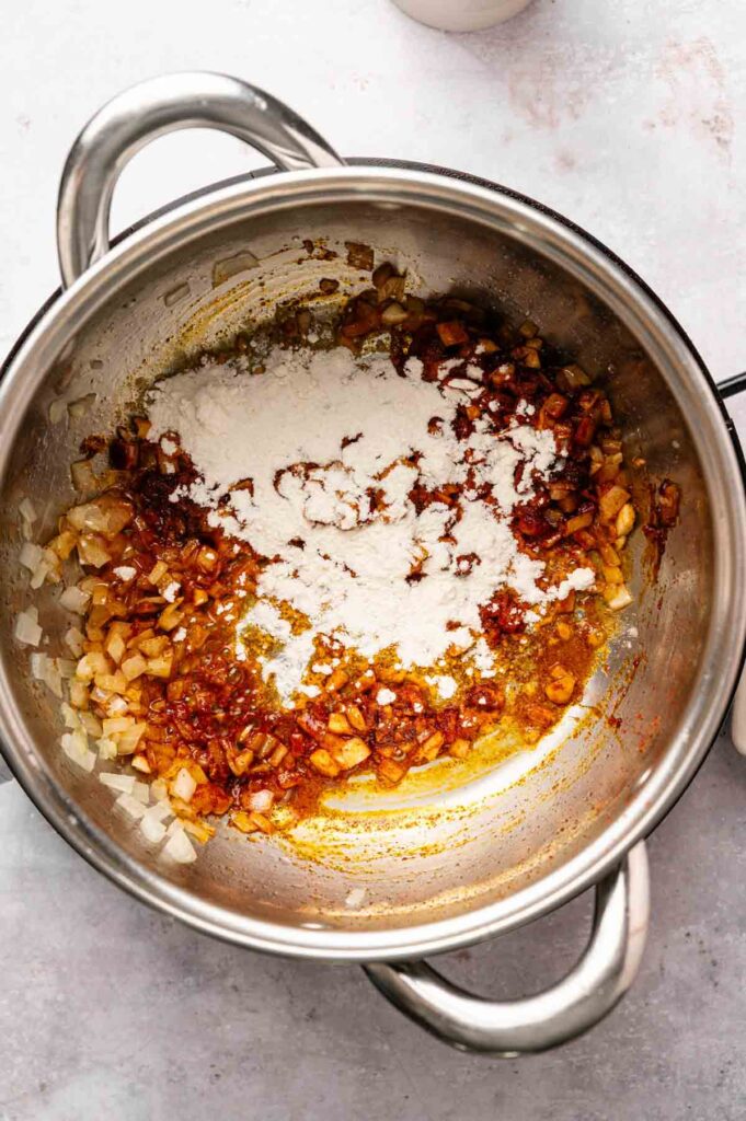 Chopped onions and seasonings are sautéed in a metal pot with flour sprinkled on top, ready to be mixed into the perfect base for Patatas Bravas.