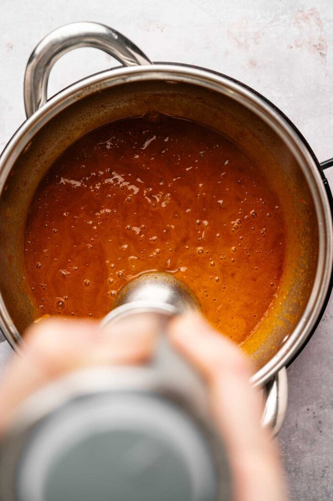 An immersion blender is being used to blend a vibrant orange Patatas Bravas sauce in a stainless steel pot on a countertop.