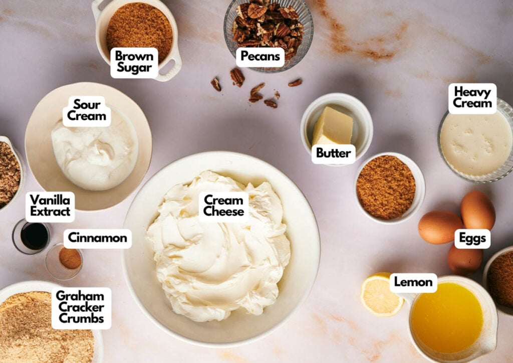 A top-down view of Pecan Pie Cheesecake ingredients, including cream cheese, eggs, lemon, butter, heavy cream, brown sugar, pecans, sour cream, vanilla extract, cinnamon, and graham cracker crumbs.