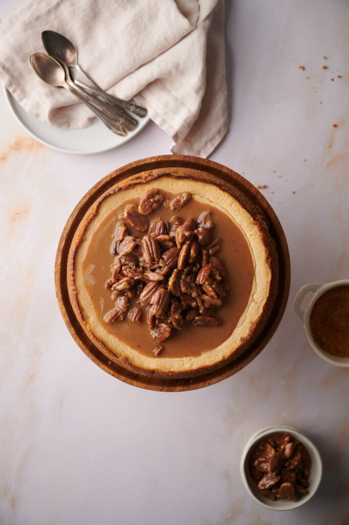 A Pecan Pie Cheesecake on a wooden stand, topped with glazed pecans, is showcased alongside a small bowl of pecans, a bowl of brown sugar, and a plate of spoons in the background.