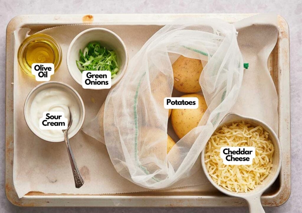 A tray with bowls containing olive oil, green onions, sour cream, cheddar cheese, and a mesh bag with potatoes&mdash;all the essentials for making delicious Loaded Potato Skins&mdash;each item clearly labeled.