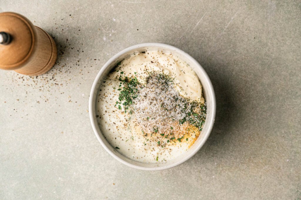 Dried herbs, salt, and pepper sit on top of mayonnaise and sour cream in a bowl, ready to be whisked together.