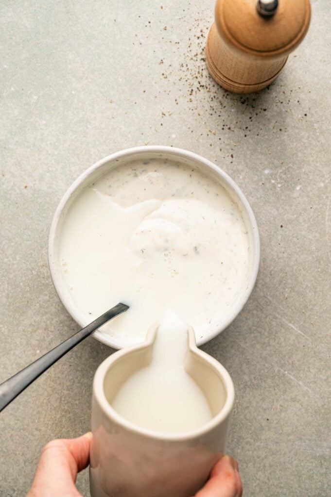 A hand pours milk from a pitcher into a bowl of creamy white ranch dressing with a spoon; a pepper grinder sits nearby on a light surface.