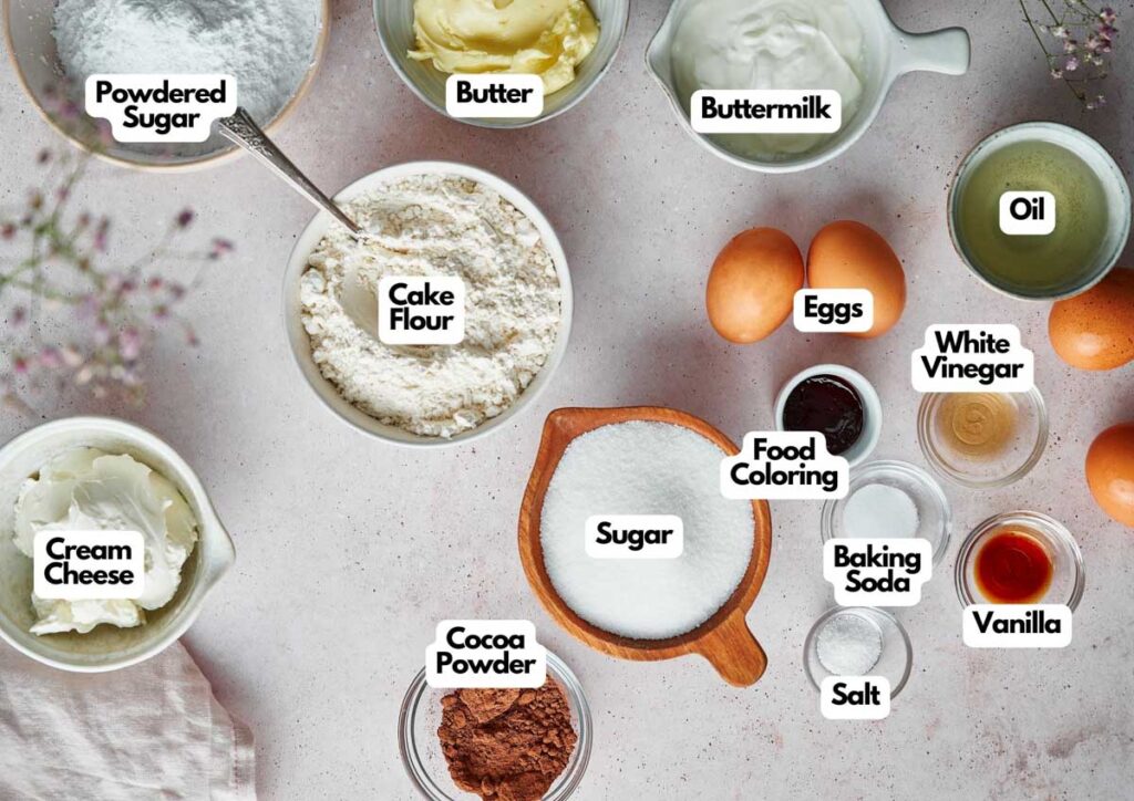 Various labeled baking ingredients for Red Velvet Cake are arranged on a tabletop, including cake flour, eggs, butter, oil, buttermilk, powdered sugar, cream cheese, sugar, cocoa powder, and several liquids.