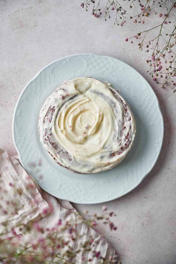 A round Red Velvet Cake with white frosting sits on a white plate, surrounded by a light cloth napkin and sprigs of small pink flowers.