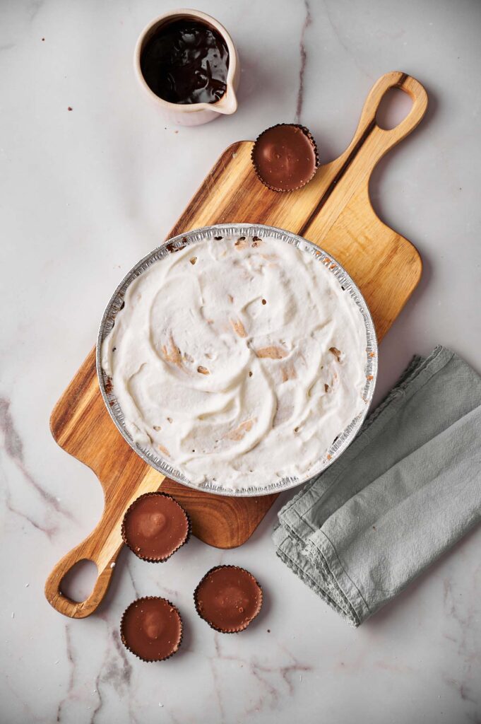 A Reese's Peanut Butter Pie with whipped topping sits on a wooden cutting board next to peanut butter cups, a small bowl of chocolate sauce, and a folded gray napkin on a marble surface.