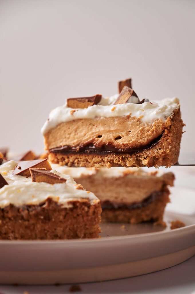 A slice of Reese's Peanut Butter Pie with a thick cookie crust, creamy layers, whipped topping, and chocolate pieces is being lifted from a plate.