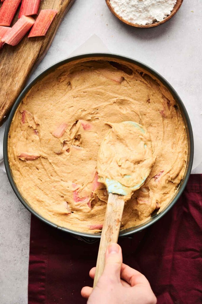 A hand spreads thick cake batter with visible rhubarb pieces in a round baking pan, preparing an Air Fryer Rhubarb Cake next to a cutting board with rhubarb and a bowl of flour.