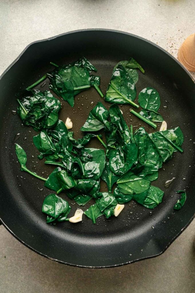 Fresh spinach and garlic cloves sauté in a skillet, wilting quickly to keep excess moisture off the pizza crust.