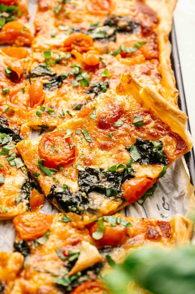 Close-up of rectangular slices of Sheet Pan Pizza topped with melted cheese, cherry tomatoes, spinach, and chopped fresh herbs on a baking sheet.