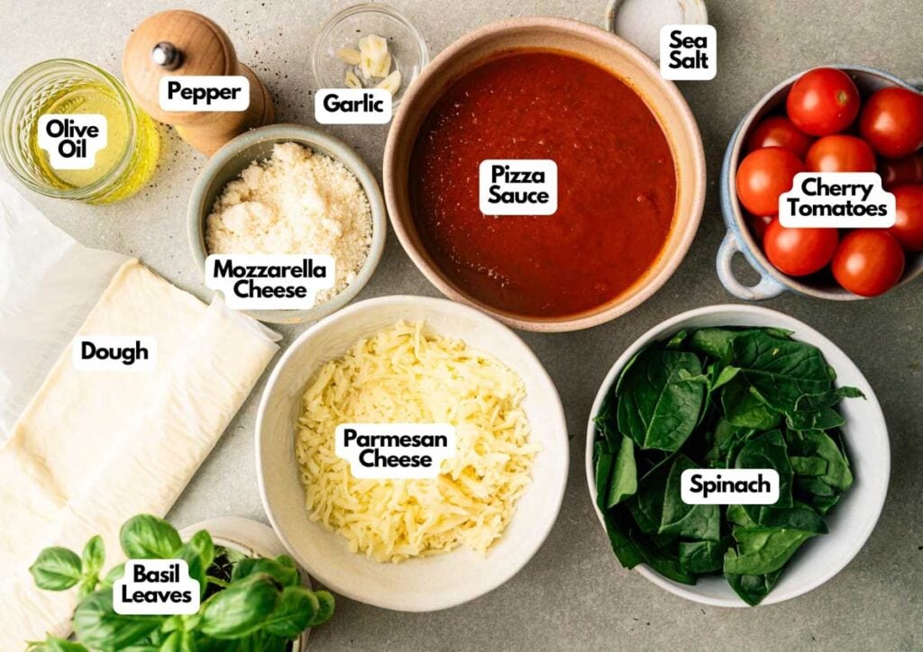 Ingredients for homemade Sheet Pan Pizza including dough, olive oil, pepper, garlic, mozzarella and parmesan cheese, pizza sauce, sea salt, cherry tomatoes, spinach, and basil leaves arranged on a counter.