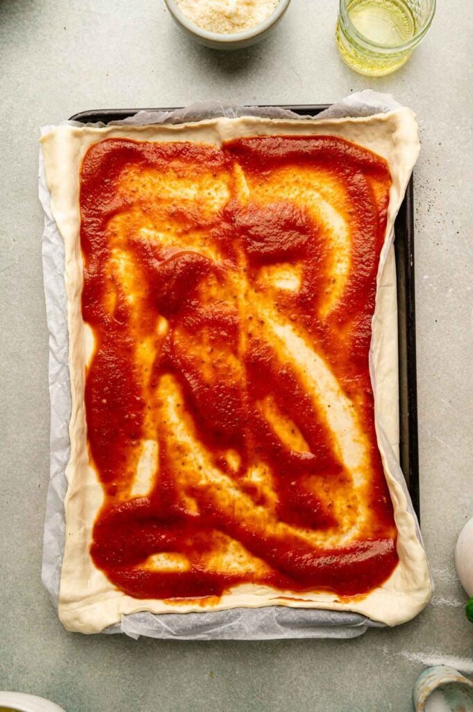 Pizza sauce is spread over stretched dough on a sheet pan, leaving a border around the edges.