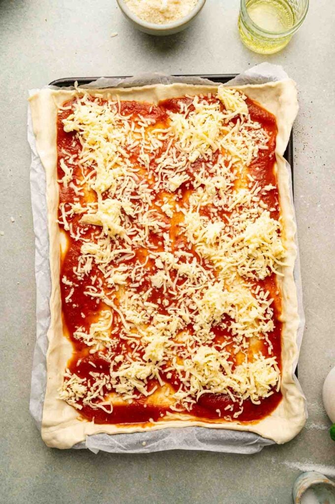 Shredded mozzarella is scattered over the sauced pizza dough, ready for the remaining toppings.