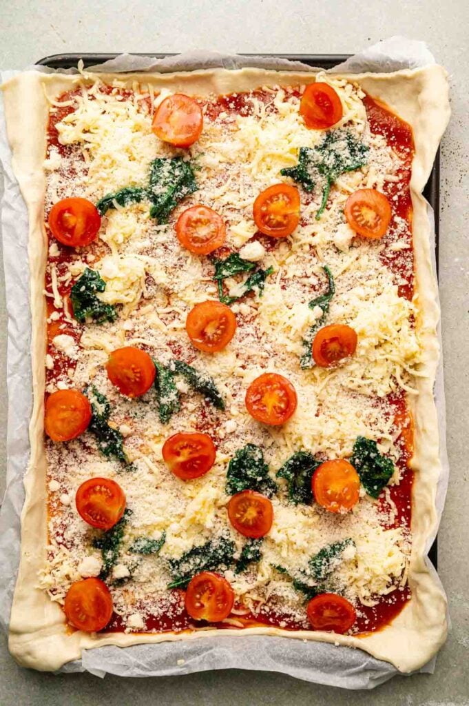Sheet Pan Pizza featuring rectangular homemade dough, tomato sauce, shredded cheese, cherry tomatoes, spinach, and grated cheese&mdash;all baked to perfection on a parchment-lined baking sheet.