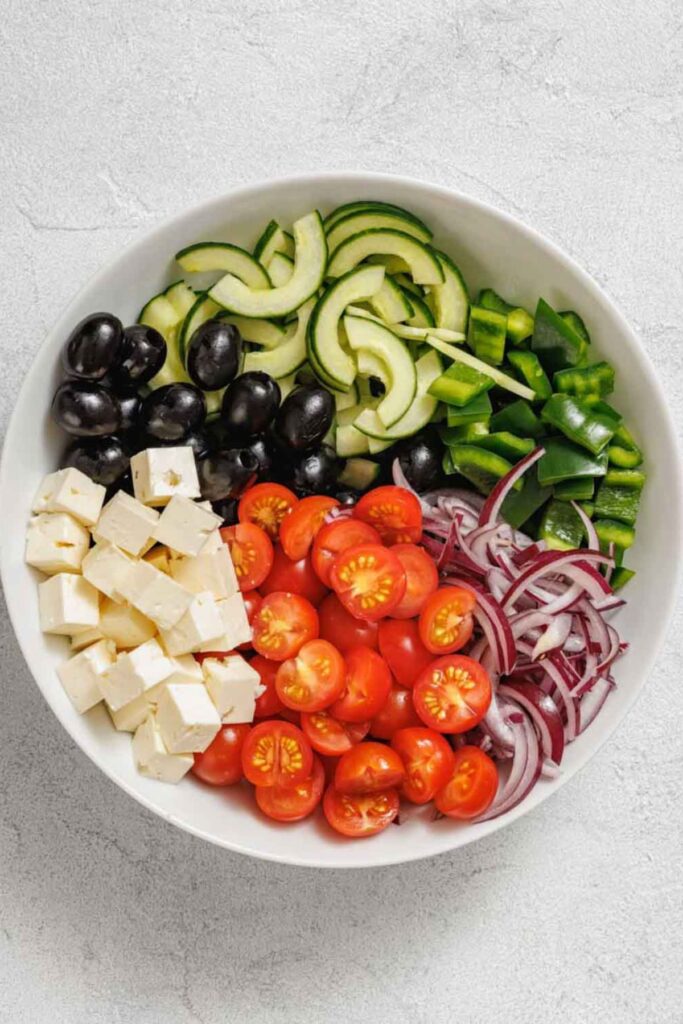 A white bowl with sliced cucumbers, green bell peppers, black olives, feta cubes, cherry tomatoes, and red onion arranged in sections—this is the essence of a Southern Farmhouse Feta & Veggie Salad.