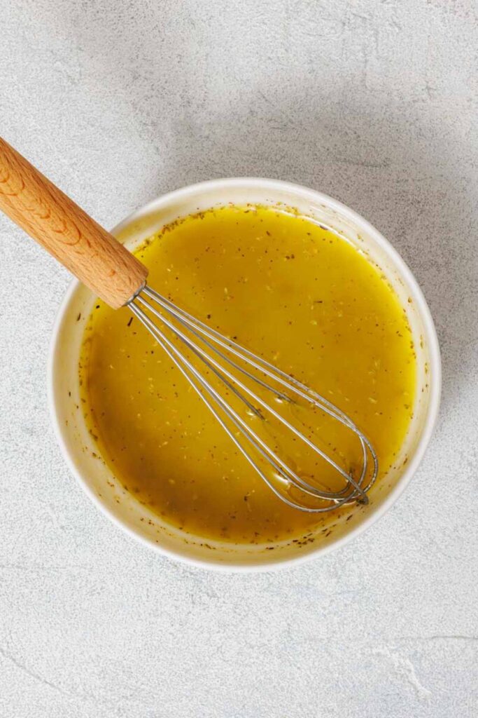 A metal whisk rests in a white bowl filled with yellow vinaigrette dressing on a light gray surface, ready to be drizzled over a Southern Farmhouse Feta & Veggie Salad.