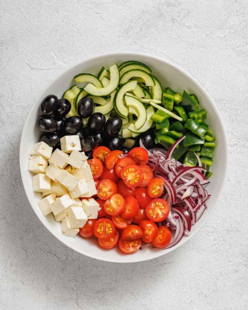 A white bowl of Southern Farmhouse Feta & Veggie Salad with sliced cucumbers, green bell peppers, black olives, cubed feta cheese, cherry tomatoes, and red onions on a light gray surface.