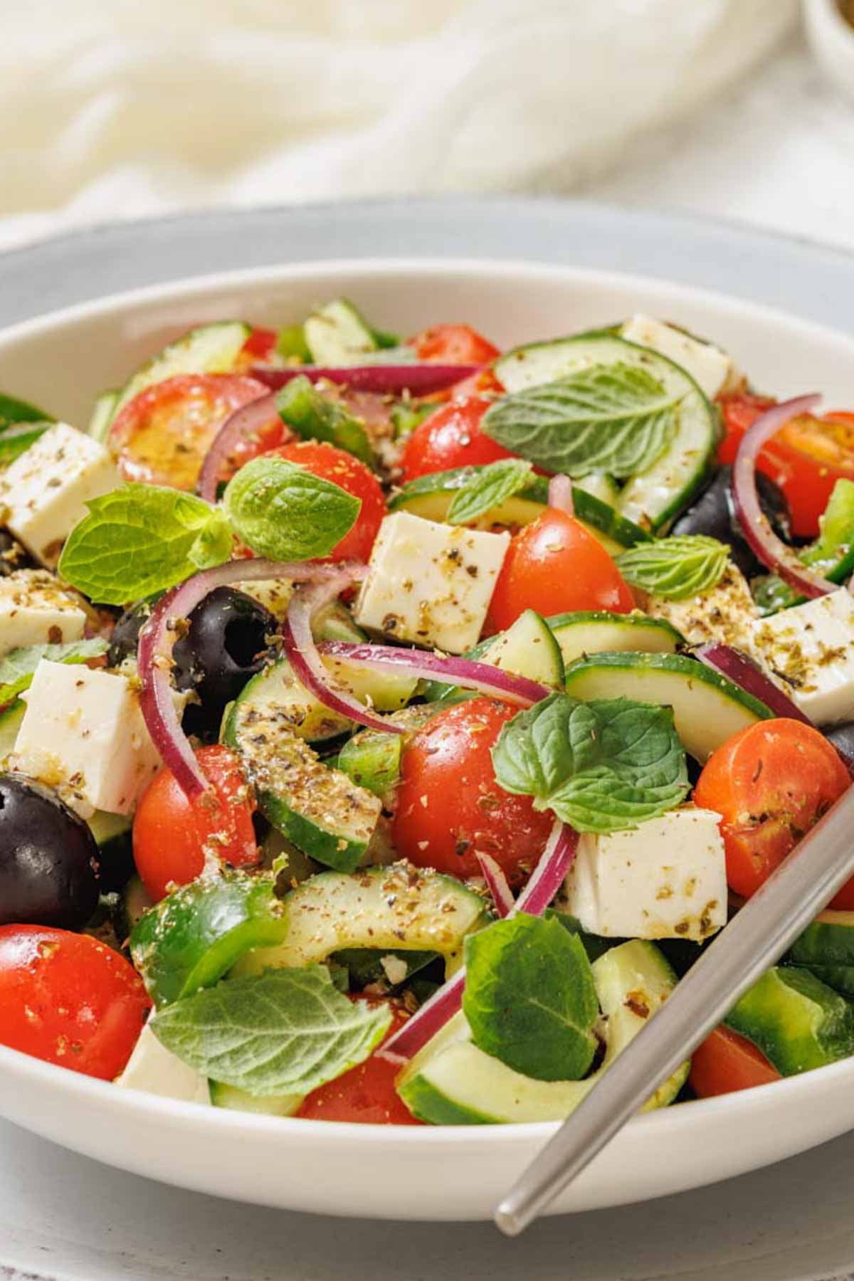 A bowl of Southern Farmhouse Feta & Veggie Salad with cherry tomatoes, cucumbers, red onions, olives, feta cheese, fresh herbs, and a fork.