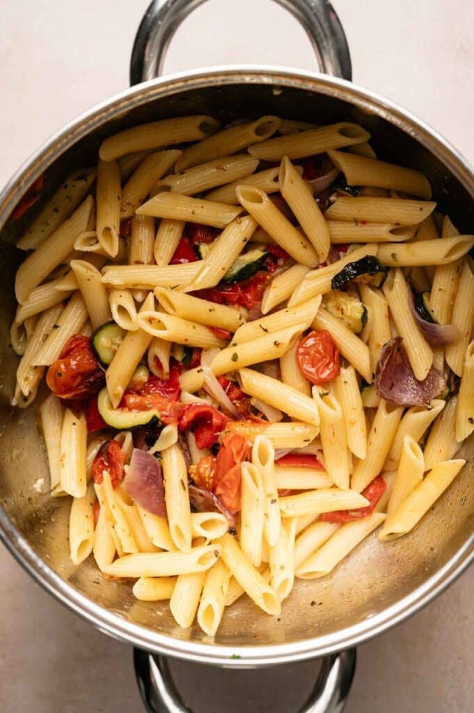 A stainless steel pot containing cooked vegetable pasta made with penne, roasted cherry tomatoes, zucchini, red onion, and herbs.