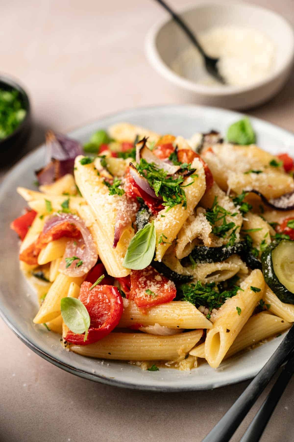 Plate of vegetable pasta featuring penne with roasted tomatoes, zucchini, and red onions, garnished with fresh herbs and grated cheese.