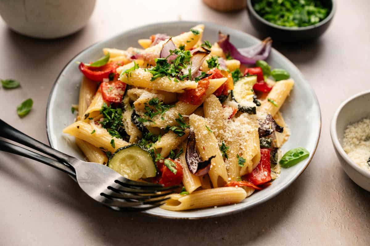 A plate of vegetable pasta featuring penne mixed with roasted vegetables, topped with grated cheese and chopped herbs, served with a fork and spoon on the side.