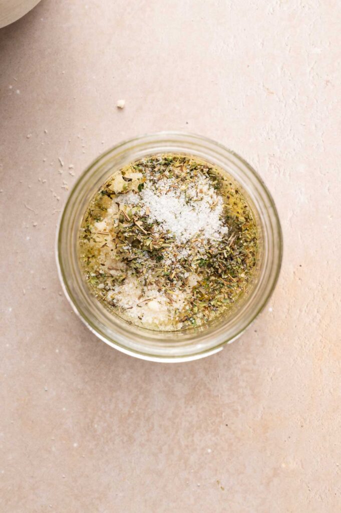 Top view of a glass jar filled with a flavorful mixture of dried herbs, grated cheese, and salt on a light-colored surface—perfect for sprinkling over your favorite vegetable pasta dishes.