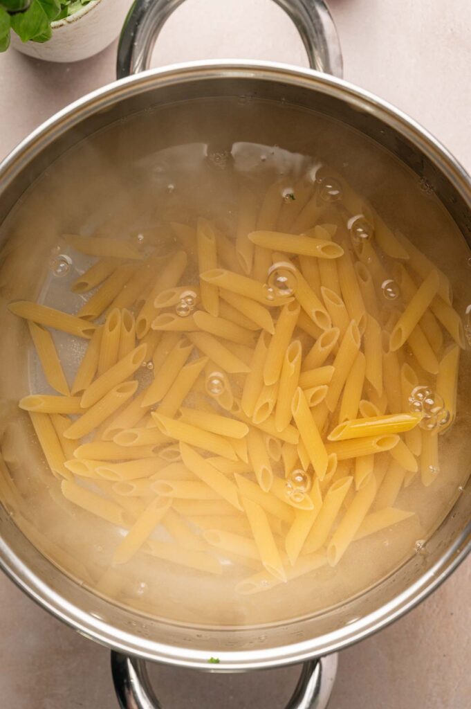 Uncooked vegetable pasta penne boiling in a stainless steel pot of water, with steam rising from the surface.
