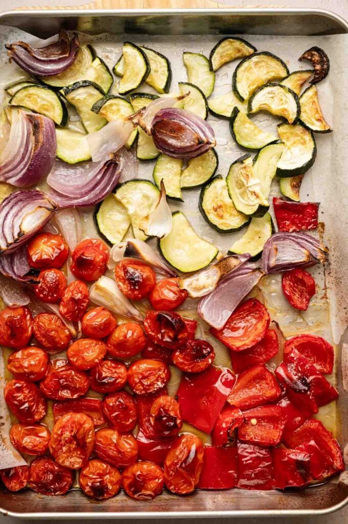A baking sheet with roasted cherry tomatoes, red bell peppers, red onions, and zucchini slices arranged in sections—perfect for tossing into your favorite vegetable pasta.