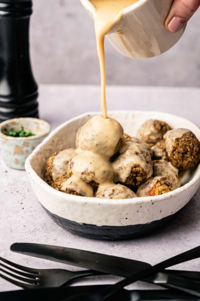 A hand pours creamy sauce over a bowl of vegetarian meatballs. Black cutlery and a small bowl of herbs are on the table nearby.