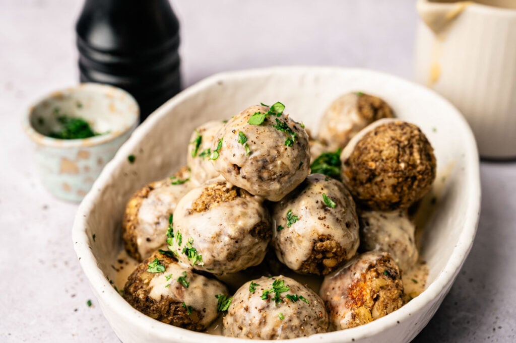 A white bowl filled with several vegetarian meatballs topped with creamy sauce and chopped herbs, with a pepper grinder and small bowl in the background.