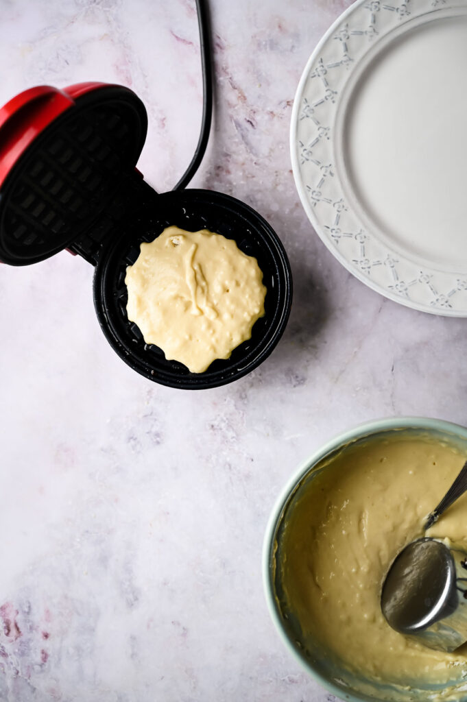 A mini waffle maker with batter inside, a bowl of batter with a spoon, and two empty plates on a light countertop—perfect for trying out your favorite waffle recipe.