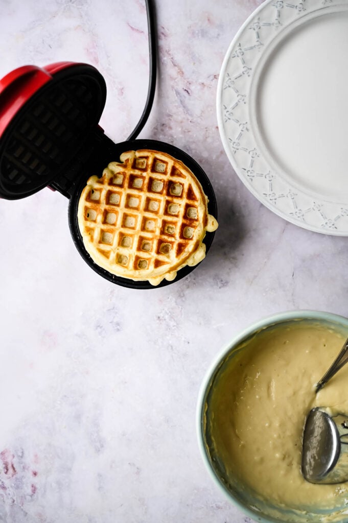 A cooked waffle in a mini waffle maker sits next to an empty plate and a bowl of waffles batter with a spoon on a light countertop.