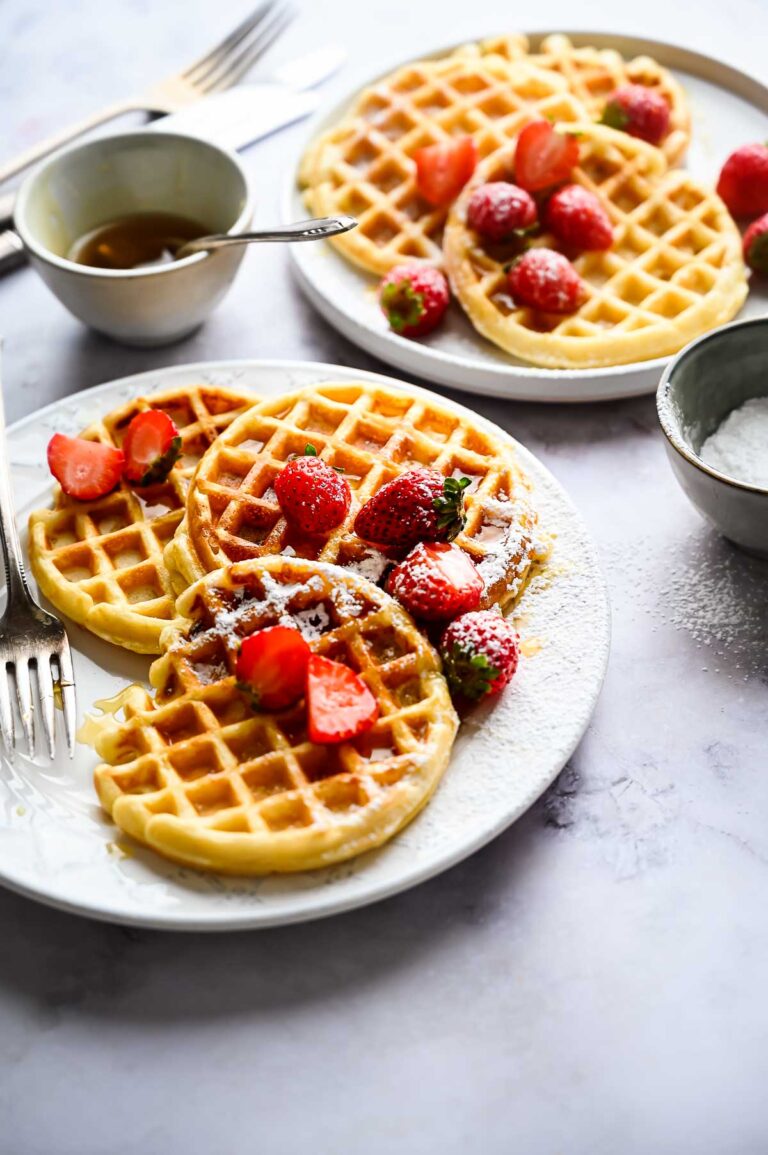 Plates of golden waffles topped with sliced strawberries and syrup, accompanied by a small bowl of syrup and a fork on a light-colored surface.